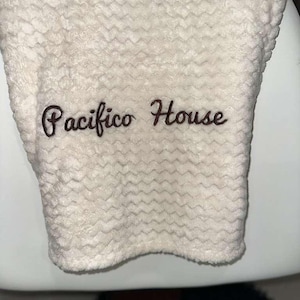 May include: Cream-colored, textured towel with the embroidered text "Pacifico House" in a dark brown script. The towel has a wavy pattern and appears soft and plush.