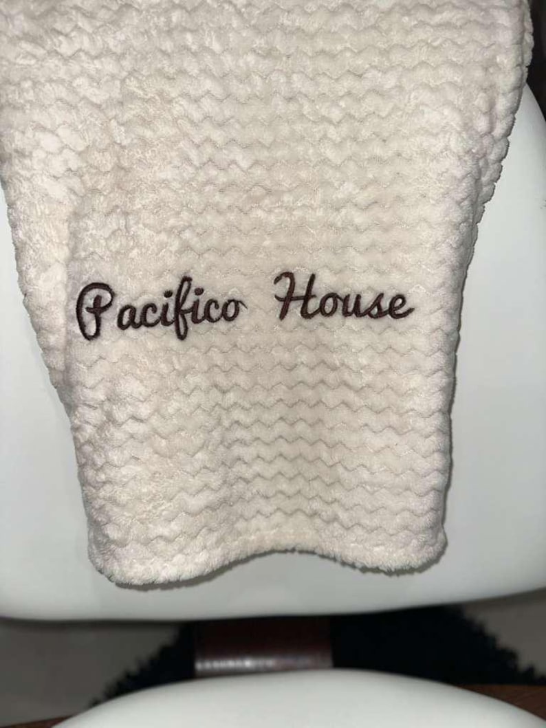 May include: Cream-colored, textured towel with the embroidered text "Pacifico House" in a dark brown script. The towel has a wavy pattern and appears soft and plush.