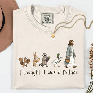 May include: A cream-colored t-shirt featuring a graphic of a squirrel with an acorn, a rabbit with bread, a raccoon with grapes, a duck with a fish, and a figure in a robe carrying a basket, with the text "I thought it was a Potluck" below.