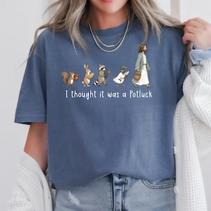 May include: A blue t-shirt featuring a graphic of a squirrel, rabbit, raccoon, duck, and a figure in robes walking in a line, each carrying food. Below the illustration, the white text reads, I thought it was a Potluck. The shirt has short sleeves.