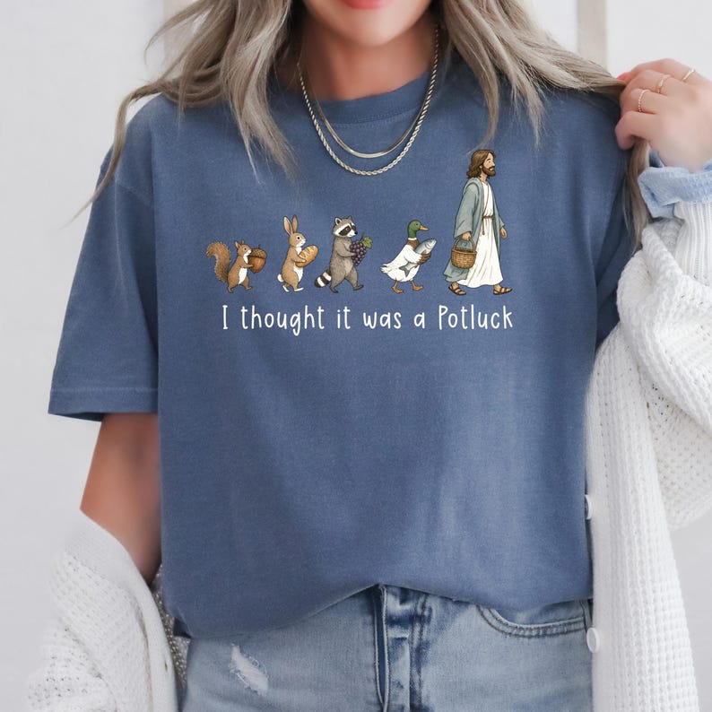 May include: A blue t-shirt featuring a graphic of a squirrel, rabbit, raccoon, duck, and a figure in robes walking in a line, each carrying food. Below the illustration, the white text reads, I thought it was a Potluck. The shirt has short sleeves.