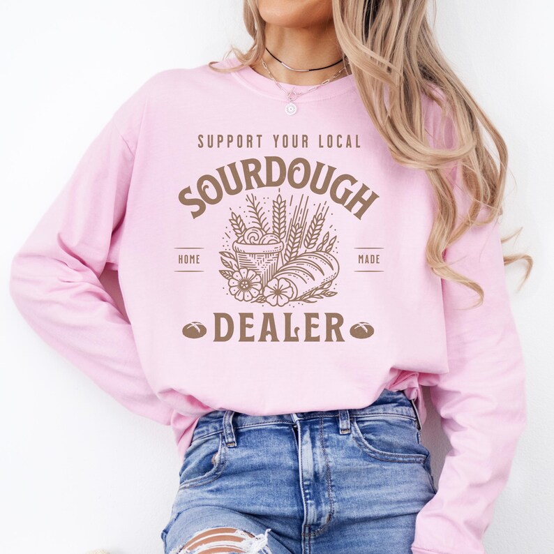 K&ouml;nnte beinhalten: Ein rosafarbenes Sweatshirt mit langen &Auml;rmeln mit dem Text "Support Your Local Sourdough Home Made Dealer" in braunen Buchstaben. Der Text ist von einer Grafik mit Weizen&auml;hren, einem Laib Brot und einem Korb mit Teig umgeben.