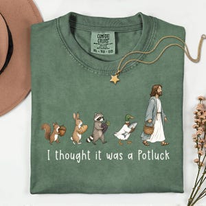 May include: A sage green t-shirt featuring a graphic of a squirrel, rabbit, raccoon, duck, and a figure in robes walking in a line. Each animal carries food, and the text reads, I thought it was a Potluck. The shirt has a Comfort Colors brand label.