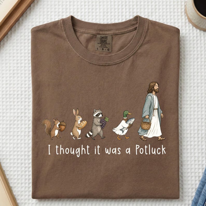 May include: A brown crewneck t-shirt featuring a graphic of a squirrel with an acorn, a rabbit with bread, a raccoon with grapes, a duck with a fish, and a figure in robes with a basket. The text reads, I thought it was a Potluck.