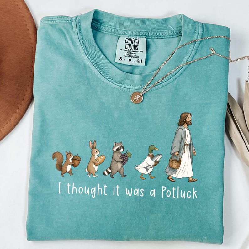 May include: A teal crewneck t-shirt featuring a graphic of a squirrel, rabbit, raccoon, duck, and a figure in robes carrying food items. The text reads I thought it was a Potluck. The shirt has a Comfort Colors label indicating size S-CH.