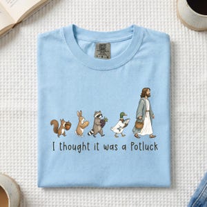 May include: A light blue t-shirt featuring a graphic of a squirrel, rabbit, raccoon, duck, and a figure in robes walking in a line, each carrying food items. Below the illustration, the text reads, I thought it was a Potluck in black font.