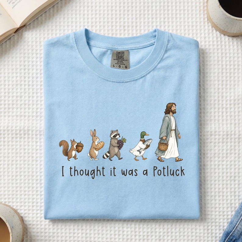 May include: A light blue t-shirt featuring a graphic of a squirrel, rabbit, raccoon, duck, and a figure in robes walking in a line, each carrying food items. Below the illustration, the text reads, I thought it was a Potluck in black font.