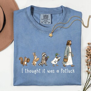 May include: A blue short-sleeve t-shirt featuring a graphic of a squirrel, rabbit, raccoon, duck, and a figure in robes walking in a line. Each carries food items like an acorn, bread, grapes, and a fish. The text reads, I thought it was a Potluck.