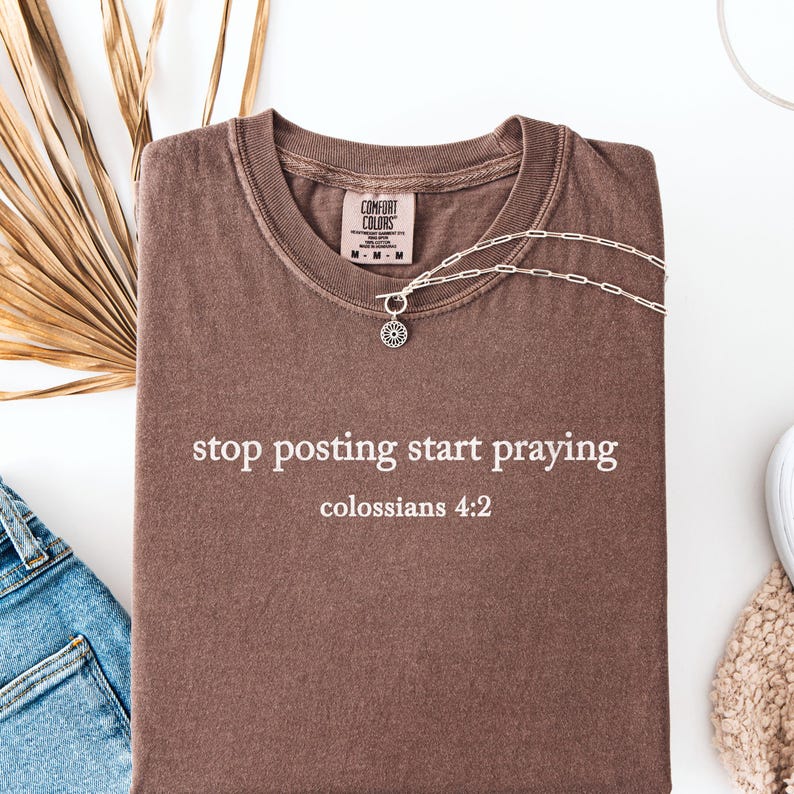 Puede incluir: Una camiseta marr&oacute;n con el texto "stop posting start praying" y "colossians 4:2" impreso en blanco.