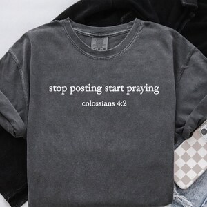 Puede incluir: Una camiseta gris oscuro con el texto "stop posting start praying" y "colossians 4:2" impreso en blanco.