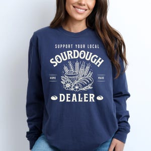 K&ouml;nnte beinhalten: Ein marineblaues Langarmshirt mit einem wei&szlig;en Grafikdesign, auf dem "Unterst&uuml;tze deinen lokalen Sauerteig-H&auml;ndler" steht. Die Grafik zeigt ein Brot, Weizen&auml;hren und Blumen.