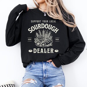 K&ouml;nnte beinhalten: Ein schwarzes Sweatshirt mit einem wei&szlig;en Grafikdesign, auf dem "Support Your Local Sourdough Home Made Dealer" steht, mit einer Zeichnung eines Laibes Brot, Weizen und einer Sch&uuml;ssel.