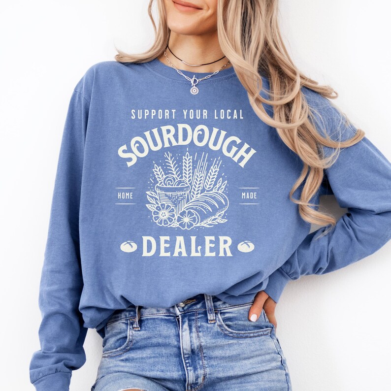 K&ouml;nnte beinhalten: Ein blaues Sweatshirt mit dem Text "Support Your Local Sourdough Home Made Dealer" in Wei&szlig;. Der Text ist von einer Grafik mit einem Brotlaib, Weizen und Blumen umgeben.