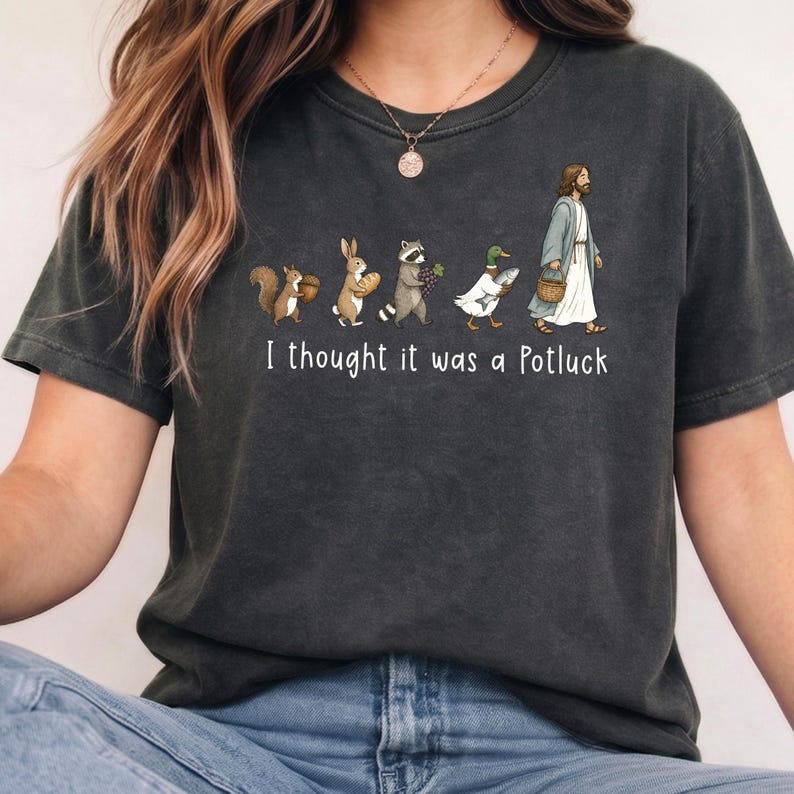 May include: A charcoal gray short-sleeve t-shirt featuring a graphic of a squirrel, rabbit, raccoon, duck, and a figure in robes walking in a line. Each carries food items. Below the graphic, the white text reads: I thought it was a Potluck.