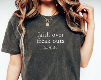 May include: Dark gray t-shirt with the text "faith over freak outs" and the reference "Isa. 41:10" in white. The shirt is a relaxed fit, with a crew neck and short sleeves. The model is wearing a gold necklace.