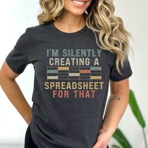 May include: A dark heather grey t-shirt with a vintage-style graphic that reads "I'm silently creating a spreadsheet for that". The graphic features a stylized spreadsheet with colorful cells.
