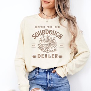 K&ouml;nnte beinhalten: Ein cremefarbener Langarmsweatshirt mit einem braunen Grafikdesign, auf dem "Support Your Local Sourdough Home Made Dealer" steht. Die Grafik zeigt ein Brot, eine Sch&uuml;ssel mit Teig und Weizen&auml;hren.
