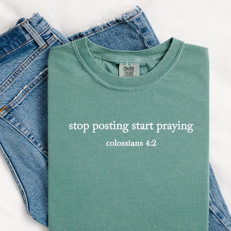 Puede incluir: Una camiseta verde azulada con el texto "stop posting start praying" y "colossians 4:2" en letras blancas. La camiseta est&aacute; colocada sobre una superficie blanca con un par de pantalones vaqueros azules.