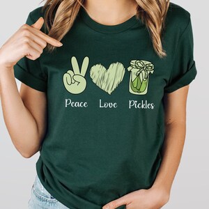 May include: A dark green t-shirt with a peace sign, a heart, and a jar of pickles. The text "Peace Love Pickles" is printed below the images.