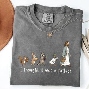 May include: A charcoal gray t-shirt featuring a graphic of a squirrel, rabbit, raccoon, duck, and a figure in robes walking in a line. Each carries food items like an acorn, bread, grapes, a fish, and a basket. The text reads, I thought it was a Potluck.