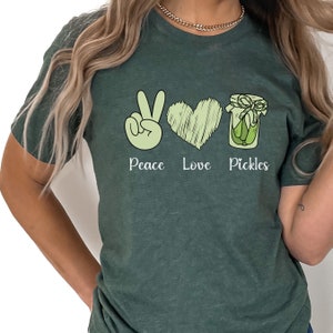 May include: A green t-shirt with a peace sign, heart, and jar of pickles. The text "Peace Love Pickles" is printed on the shirt.