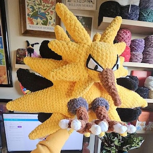 Pode incluir: Um grande peluche amarelo de crochê feito à mão de uma criatura semelhante a um pássaro com um longo bico castanho e olhos pretos e brancos. O peluche tem penas amarelas pontiagudas e pés castanhos e brancos.