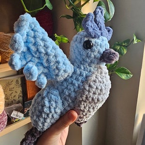 May include: A crocheted blue and gray bird plush toy with a crown on its head. The bird has black eyes and a purple beak.