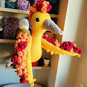 Könnte beinhalten: Ein gehäkeltes Vogelspielzeug mit gelbem Körper, orangefarbenen und roten Akzenten und einem langen Schnabel. Der Vogel sitzt auf einem Regal mit Wollknäueln im Hintergrund.