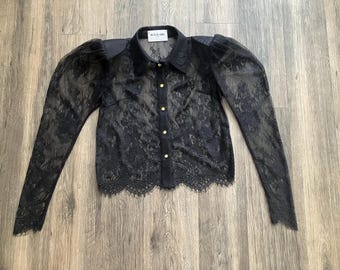May include: Black lace blouse with long sleeves and a button-down front. The blouse features a collared neckline and gold-colored buttons. The sleeves have a sheer, puffy design. The bottom hem is scalloped. The label reads "NATALIA FORS".