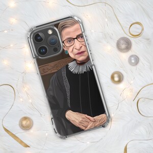 May include: A clear phone case with a portrait of Ruth Bader Ginsburg. The case features a black and white image of Ginsburg in her judicial robes, with a white lace collar.