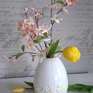 Könnte beinhalten: Eine weiße, eiförmige Vase mit Gänseblümchenmuster hält rosa und weiße Blumen und grüne Blätter. Gelbe Tulpen sind um die Vase platziert. Der Hintergrund ist eine hellgraue Wand mit Schrift.