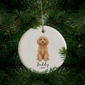 May include: A round white ceramic ornament with a marble texture, featuring a detailed illustration of a golden-brown curly-haired dog sitting in the center. Below the dog, the name Buddy and the year 20xx are printed in black text.