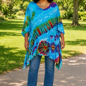 May include: A bright blue tie-dye tunic featuring a V-neckline, short wide sleeves, and an asymmetrical hemline with vibrant red, orange, and purple circular patterns, paired with medium-wash blue denim jeans and tan open-toe sandals.