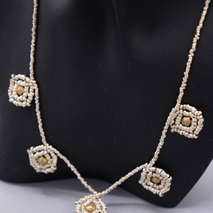 Könnte beinhalten: Silberne Perlenkette mit quadratischen, goldfarbenen Akzenten in der Mitte. Die Halskette wird auf einer schwarzen Schaufensterpuppe präsentiert. Die Halskette hat ein zartes, aufwendiges Design mit einer Reihe von quadratischen Formen.