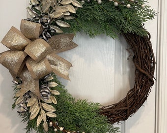 May include: A decorative wreath featuring a circular grapevine base adorned with green faux evergreen branches, gold glittery leaves, and a large gold bow. White and silver berries add a festive touch to this holiday decoration.