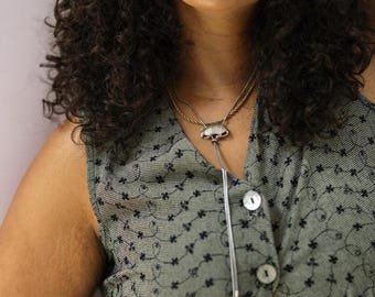 May include: A silver-toned bolo tie featuring a small, metallic animal-shaped slide, worn over a sleeveless olive green vest with a black embroidered floral pattern and white buttons. The bolo tie has two long, thin silver-toned chains.