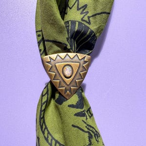 May include: A bronze-toned metal scarf slide with a triangular shape and a central opal-like stone. The slide is adorned with a geometric pattern and holds a green scarf with black illustrations. The scarf is knotted, showcasing the slide's function.