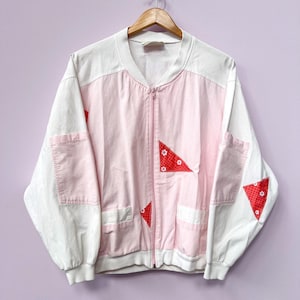 May include: A vintage bomber jacket in white and pink, with a zip-up front and pockets. The jacket features red triangle patches with white floral designs. The sleeves and collar are white, while the body is pink. The jacket is hanging on a wooden hanger.