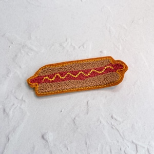 May include: Embroidered hot dog patch with a brown bun, red filling, and yellow zigzag mustard design. The patch has a brown border and is set against a white textured background. This food-themed patch is suitable for jackets, bags, or other fabric items.