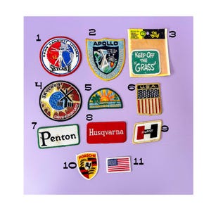 May include: A collection of vintage embroidered patches on a purple background. Patches include the American flag, "U.S.A.", "Apollo", "Husqvarna", "Penton", "Porsche", and "Keep Off The Grass".