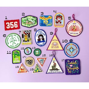 May include: A collection of colorful embroidered patches with various designs and text. Patches include a red "356" patch, a "Happy Birthday" patch, a tiger graduate patch, and a compass patch. The patches are of various shapes and sizes.