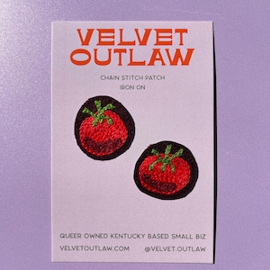 May include: A white card with the text "VELVET OUTLAW" in orange letters. Below the text are two embroidered tomato patches with red tomatoes and green stems. The card also reads "CHAIN STITCH PATCH IRON ON".