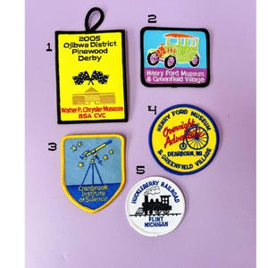 May include: A collection of five embroidered patches with various designs and text. The patches feature a yellow rectangle with "2005 Ojibwa District Pinewood Derby", a green car, a telescope, a bicycle, and a train. The patches are in various colors.