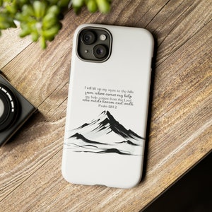 May include: White phone case featuring a black and white mountain illustration with the text "I will lift up my eyes to the hills from where comes my help...Psalm 121:1-2". The case is on a wooden surface.