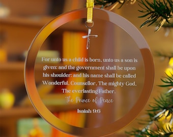 May include: A clear, circular Christmas ornament with a gold ribbon. The ornament displays a cross and text from Isaiah 9:6, including "For unto us a child is born..." and "The Prince of Peace."