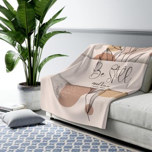 May include: A soft, beige throw blanket with a floral design and the text "Be Still and Know" in black script. The blanket is draped over a white couch.