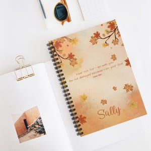 Puede incluir: Cuaderno de espiral con una cubierta con temática otoñal. La cubierta presenta hojas de otoño en tonos naranja, amarillo y marrón, con el nombre "Sally" y un versículo bíblico. El cuaderno está abierto sobre una superficie blanca con otros artículos de papelería.
