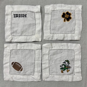 Könnte beinhalten: Vier weiße Leinenuntersetzer mit gestickten Designs. Einer hat das Wort "IRISH" in Schwarz gestickt. Ein anderer zeigt ein Kleeblatt mit "ND" in Gold und Marine. Der dritte hat einen braunen Football und der letzte einen Kobold.