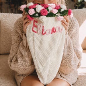 May include: A cream-colored, cone-shaped crochet bouquet holder featuring the name Eliana embroidered in pink yarn. The top is filled with a cluster of small, round, crocheted flowers in shades of light pink, dark pink, and white with green leaves.