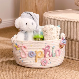 Op de afbeelding: Een crèmekleurige touwmand met de naam "Sophia" in kleurrijke letters. De mand bevat een teddybeer, een babyfles en luiers. De mand is versierd met kleurrijke bloemen en stippen. Een leuk cadeau voor een babyshower.